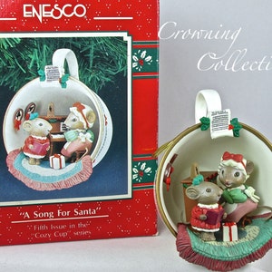 May include: A Christmas ornament shaped like a teacup, featuring two mice singing and playing a piano. The ornament has a red and white color scheme, with a gold rim and a small gift box. The text "A Song For Santa" is visible.