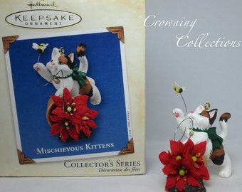 Könnte beinhalten: Hallmark Keepsake Ornament, Mischievous Kittens Collector's Series, zeigt eine weiße, braune und schwarze Katze mit grünem Band, die eine Biene jagt, die auf einem roten Weihnachtsstern steht.