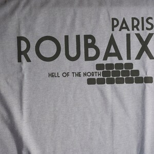 May include: A grey t-shirt with black text that reads "Paris Roubaix Hell of the North". The text is in a bold, blocky font. Below the text is a graphic of a brick wall.