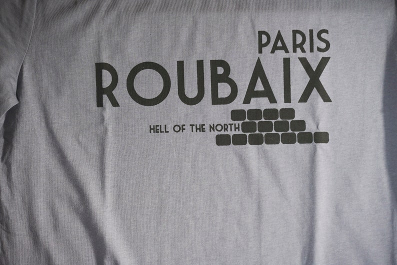 May include: A grey t-shirt with black text that reads "Paris Roubaix Hell of the North". The text is in a bold, blocky font. Below the text is a graphic of a brick wall.