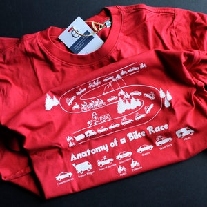 May include: Red t-shirt with a white graphic of a winding road with various vehicles and the text "Anatomy of a Bike Race". The vehicles include a team car, medic, police, TV photo, journalists, guests, brown wagon, medical service, and commissaries.