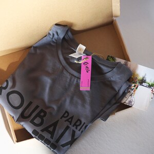 May include: A grey t-shirt with the text "Paris Roubaix" printed on the front. The shirt is folded and placed in a cardboard box with a pink tag that says "Le Velo".
