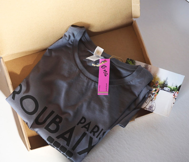 May include: A grey t-shirt with the text "Paris Roubaix" printed on the front. The shirt is folded and placed in a cardboard box with a pink tag that says "Le Velo".