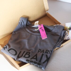 May include: A grey t-shirt with the text "DUBAI PARIS" printed on it. The shirt is folded and placed in a cardboard box with a pink tag that says "Cyclos".