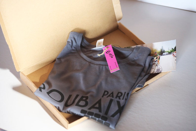 May include: A grey t-shirt with the text "DUBAI PARIS" printed on it. The shirt is folded and placed in a cardboard box with a pink tag that says "Cyclos".