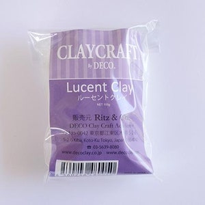 May include: A purple package of Lucent Clay, a modeling clay product by Deco Clay Craft Academy. The package is labeled with the text "Lucent Clay" in English and Japanese, and "Ritz & Deco Clay Craft Academy" in English. The package is 100g.