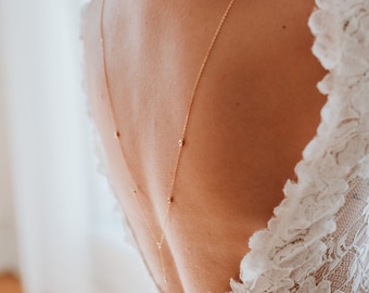 May include: A white lace wedding dress with a delicate back detail featuring a gold chain with small clear stones.