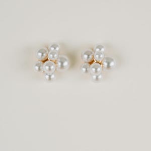 May include: A pair of pearl cluster earrings. Each earring features a gold-toned base with multiple faux pearls arranged in a floral-like design. The earrings are set against a neutral background.