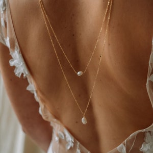 May include: A back-length gold chain necklace with two pearl pendants. The necklace is worn by a person with their back facing the camera.