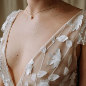 May include: A woman wearing a sheer, nude-colored dress with white floral embroidery. The dress has a deep V-neckline and a delicate pearl necklace.