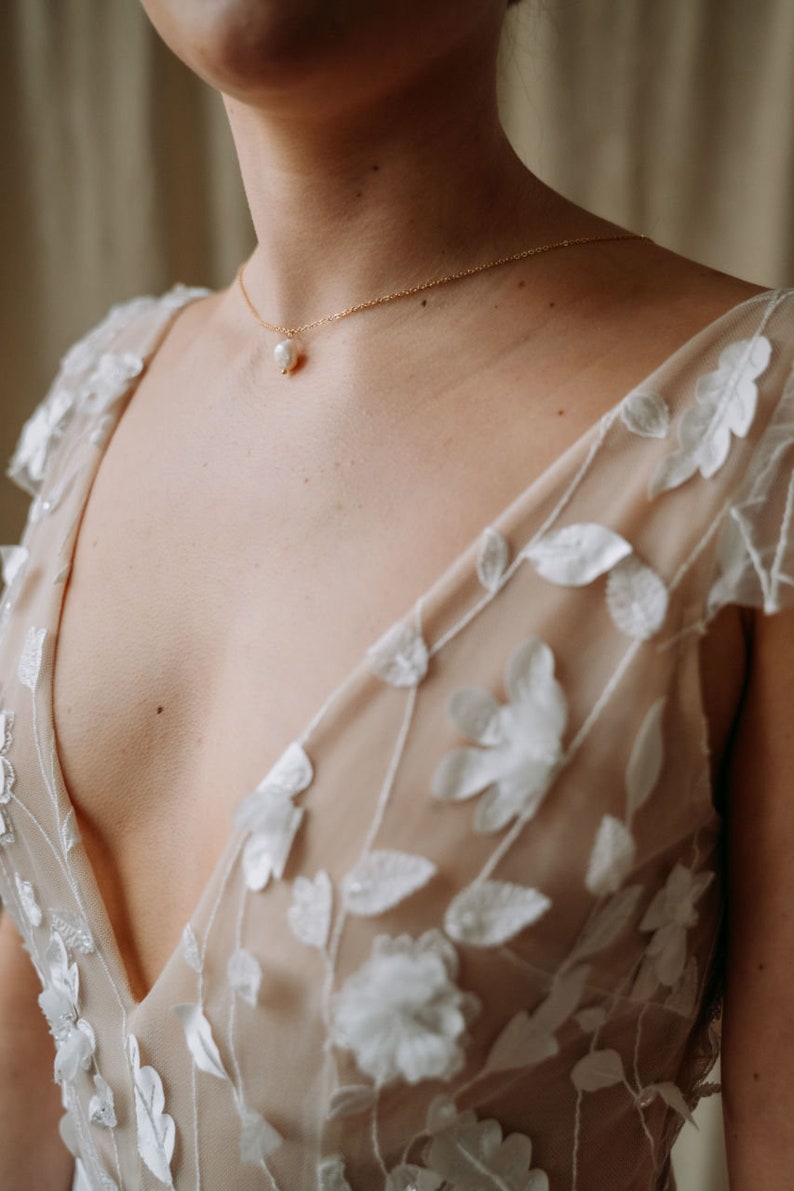 May include: A woman wearing a sheer, nude-colored dress with white floral embroidery. The dress has a deep V-neckline and a delicate pearl necklace.