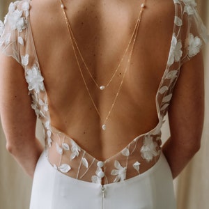 May include: A white bridal gown with a low back and delicate floral lace detailing. The gown features a back-length pearl necklace with three pearls.