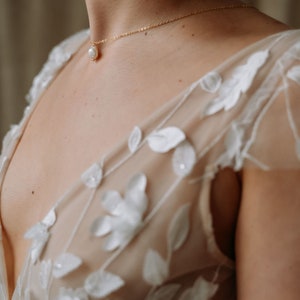 May include: A woman wearing a white sheer dress with floral embroidery. She is wearing a delicate gold necklace with a single pearl pendant.