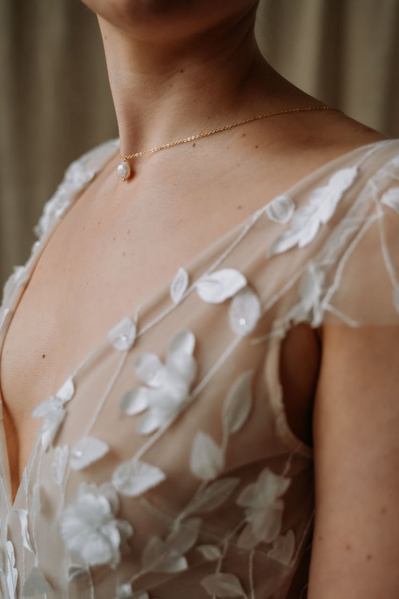 May include: A woman wearing a white sheer dress with floral embroidery. She is wearing a delicate gold necklace with a single pearl pendant.