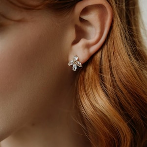 May include: A close-up of an ear with a small, gold-coloured stud earring. The earring has a cluster of clear, sparkling crystals in a floral design. The person has long, auburn hair.