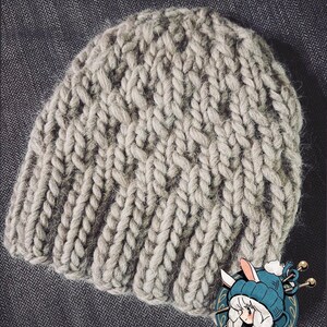 May include: A light grey knitted beanie hat with a ribbed brim and a textured, cable-knit design. The hat is made of thick yarn and appears soft and warm. The words "Knitting Pattern" are written in cursive at the bottom of the image. A cartoon image is in the bottom right corner.