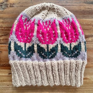 Peut inclure: Un bonnet en tricot beige orné d'un motif floral de trois tulipes roses avec des feuilles vertes et des accents gris. Le bonnet a un bord côtelé et est fait de fil doux et texturé.