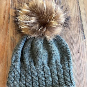 Puede incluir: Gorro de punto verde con un diseño de trenzas texturizado. Tiene un gran pompón con tonos marrones, beige y negros. El gorro está sobre una superficie de madera.