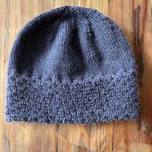 Może przedstawiać: Granatowa dzianinowa czapka z teksturowanym brzegiem. Czapka wykonana jest z miękkiego, dzianinowego materiału i ma za zadanie utrzymywać ciepło głowy. Czapka jest w jednolitym kolorze i nadaje się na zimną pogodę.