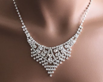 May include: A silver rhinestone necklace with a cascading design. The necklace features a large, ornate pendant with a teardrop shape.