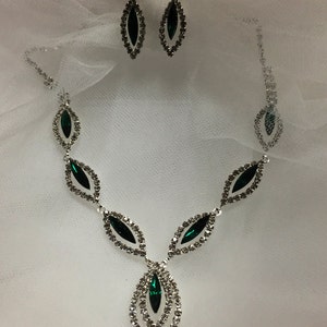 May include: A silver necklace and earrings set with emerald green stones. The necklace features a teardrop design with multiple layers of clear rhinestones.
