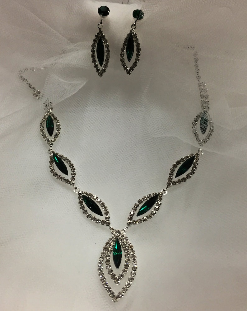 May include: A silver necklace and earrings set with emerald green stones. The necklace features a teardrop design with multiple layers of clear rhinestones.