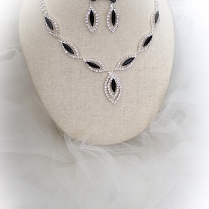 May include: A silver necklace and earrings set with black and clear rhinestones. The necklace features a teardrop pendant with a black stone surrounded by clear stones. The earrings are simple teardrop shapes with black stones.