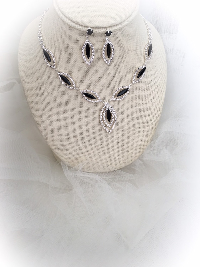 May include: A silver necklace and earrings set with black and clear rhinestones. The necklace features a teardrop pendant with a black stone surrounded by clear stones. The earrings are simple teardrop shapes with black stones.