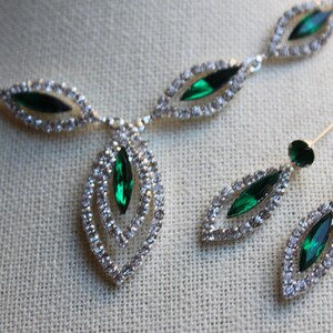 May include: A silver necklace and earrings set with emerald green stones and clear rhinestones. The necklace has a pendant with a double-layered design.