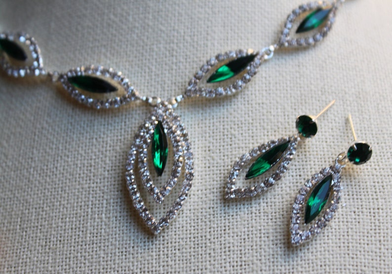 May include: A silver necklace and earrings set with emerald green stones and clear rhinestones. The necklace has a pendant with a double-layered design.
