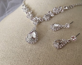 May include: A silver necklace with a teardrop-shaped pendant and matching earrings. The necklace and earrings are adorned with clear rhinestones.