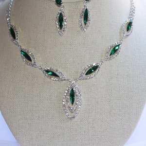 May include: A silver necklace with a teardrop pendant and emerald green rhinestones. The necklace is paired with matching earrings.