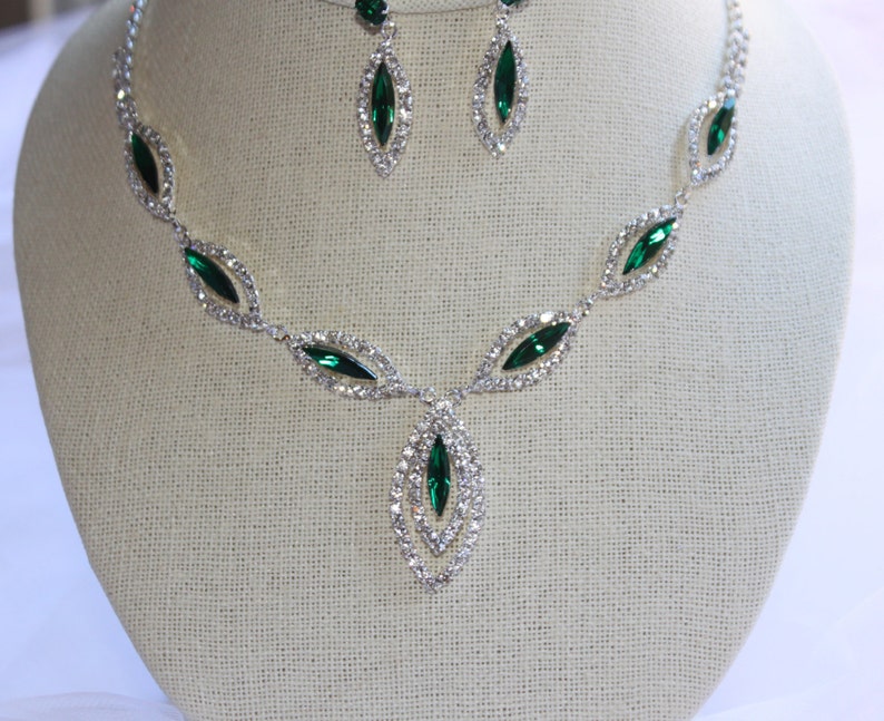 May include: A silver necklace with a teardrop pendant and emerald green rhinestones. The necklace is paired with matching earrings.