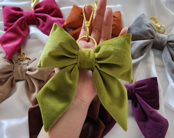 Peut inclure: Une collection de porte-clés nœuds en velours de différentes couleurs, notamment vert olive, magenta, marron, gris et orange. Chaque nœud est doté d'un fermoir mousqueton et d'un anneau en métal doré, présentant une texture douce et veloutée.