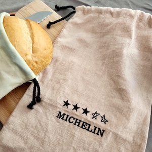 May include: A beige linen bread bag with a black drawstring, next to a matching bag with the word "MICHELIN" and five stars embroidered in black. A loaf of bread is partially visible in a cream-colored bag. A wooden cutting board and a bread knife are in the background.