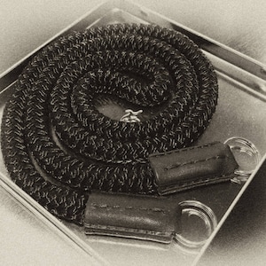 Pode incluir: Uma correia de câmera trançada preta com ferragens de metal prateado e duas correias de couro.