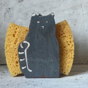 May include: A gray ceramic cat-shaped sponge holder with white accents, holding a yellow sponge. The cat has a simple design with a raised paw. The sponge is a bright yellow color, and the holder is on a white surface.