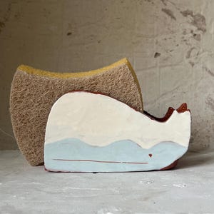 Könnte beinhalten: Ein keramischer Wal-Schwammhalter mit weißem Körper, hellblauen Wellen und braunem Sockel. Ein Naturschwamm befindet sich hinter dem Halter. Der Wal hat ein kleines rotes Herzdetail.