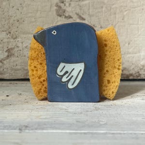 May include: A blue ceramic bird-shaped sponge holder with a white wing design and a black beak. Two yellow sponges are attached to the sides. The holder is on a white wooden surface.