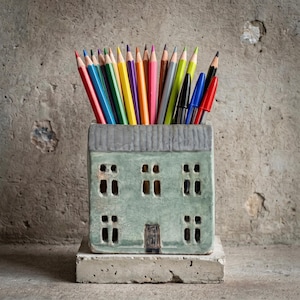Puede incluir: Un portalápices de cerámica en forma de casa de color verde claro con un techo gris y recortes rectangulares para ventanas y una puerta. El soporte está lleno de una variedad de lápices afilados de colores y varios bolígrafos rojos, azules y negros.