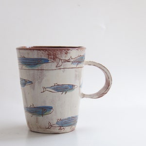 May include: A white ceramic mug with a brown rim and a handle. The mug is decorated with a blue and brown fish design.