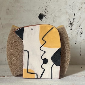 May include: A decorative ceramic sponge holder in the shape of a dog. The dog has a white face with yellow and black accents, and black line art. The holder is paired with a natural fiber sponge.