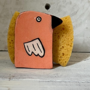 Może przedstawiać: Ceramiczny uchwyt na gąbkę w kształcie ptaka w jasnym brzoskwiniowym kolorze z czarnym dziobem i białym wzorem skrzydła. Uchwyt ma po bokach żółtą gąbkę. Ptak ma jedno czarne oko. Przeznaczony do użytku w kuchni.