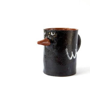 May include: A black ceramic mug with a brown rim and a painted face. The mug has a brown beak and white eyes.