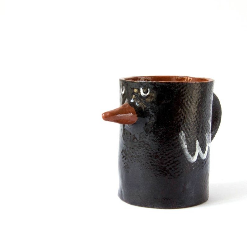 May include: A black ceramic mug with a brown rim and a painted face. The mug has a brown beak and white eyes.