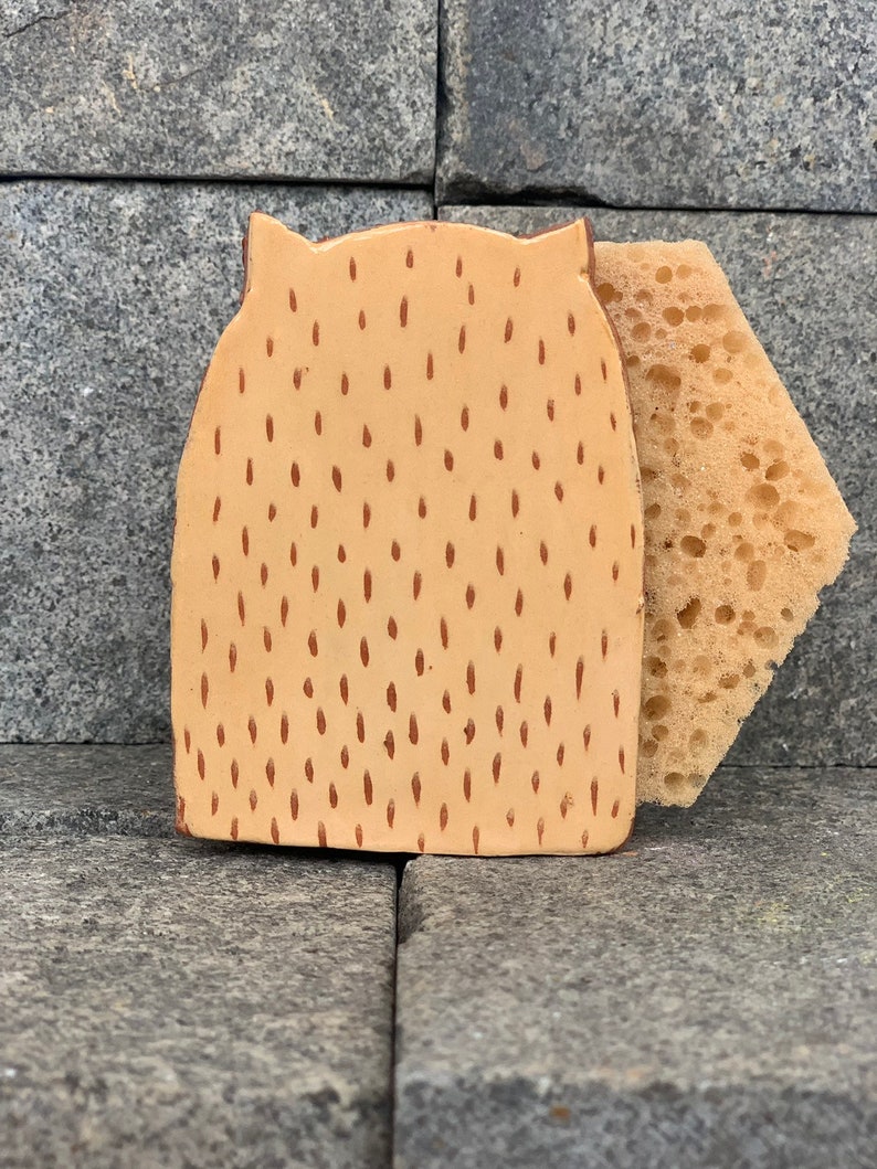 May include: A ceramic dish sponge holder shaped like an owl with a brown and tan speckled pattern. The holder has a small sponge attached to the side.
