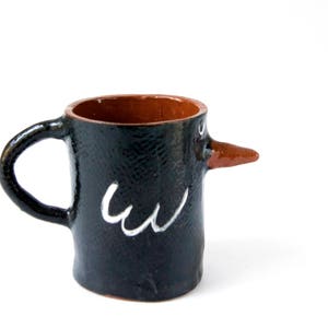 May include: A black ceramic mug with a brown clay beak and white painted details. The mug is shaped like a bird with a handle.