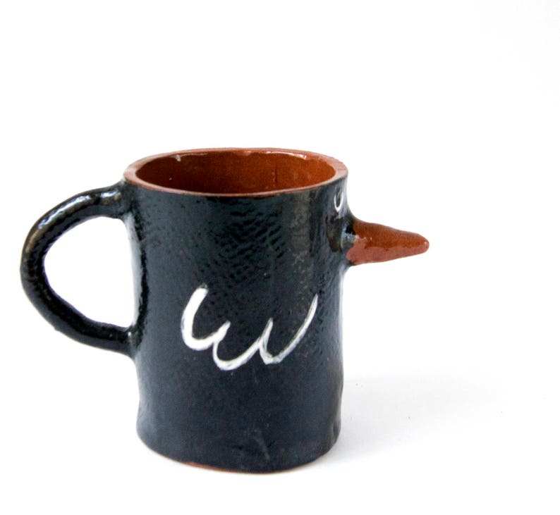May include: A black ceramic mug with a brown clay beak and white painted details. The mug is shaped like a bird with a handle.
