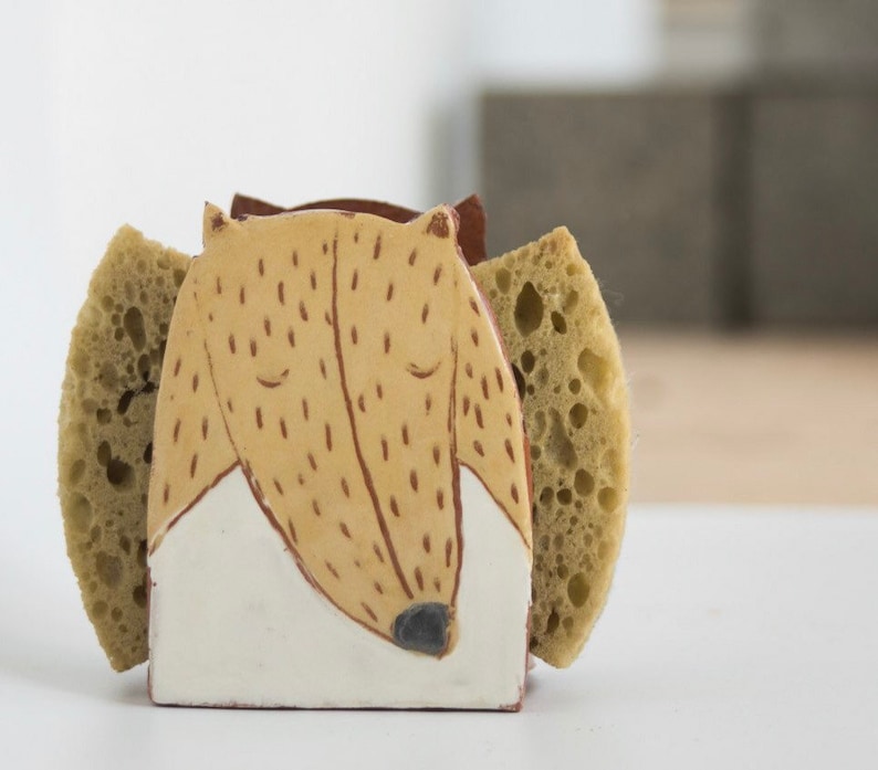 May include: Ceramic dish soap holder shaped like a dog with brown and white fur. The holder has two green sponges on either side.