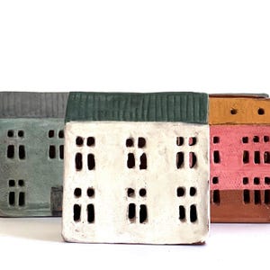 Op de afbeelding: Drie miniatuurkeramische huizen met verschillende gekleurde daken en raamuitsparingen. Het huis links is grijs, het middelste huis is wit met een groen dak en het huis rechts is bruin met een roze en geel dak.